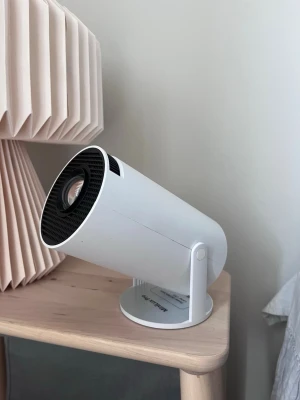 Vit kompakt projektor på stativ - Säljer en stilren, vit projektor med rund form och justerbart stativ. Projektorn har HDMI- och USB-port samt ventilationsgaller på baksidan. Perfekt för filmkvällar eller gaming och smidig att placera i hemmet tack vare sin kompakta design.