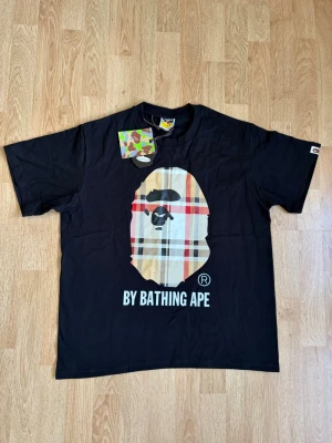 Svart BAPE t-shirt med rutig logga - Cool svart t-shirt från BAPE med den klassiska apansiluetten i beige, röd och svart rutig design på bröstet. Trycket 'BY BATHING APE' under loggan. Rund hals och korta ärmar. Perfekt för dig som gillar streetwear. Helt ny och aldrig använd T-shirt som är extremt snygg. Den passar alla årstider då den är något tjockare och perfekt för sommaren eller att ha under en hoodie i vinter. Skriv vid frågor eller funderingar!