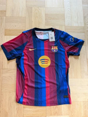 FC Barcelona hemmatröja 26/27 Player Edition - Säljer en helt ny Barca hemmatröja player edition från Nike för säsongen 2026/2027 i blått och rött med guldfärgade detaljer. Tröjan har klubbmärke, Spotify-logga och Nike Swoosh på bröstet samt korta ärmar och V-ringning. Tillverkad i lätt och ventilerande material, perfekt för fotboll. Pris kan diskuteras vid snabb affär.