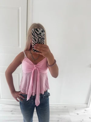Ljusrosa linnetopp med knyt - Söt ljusrosa linnetopp med smala axelband och knyt framtill. Toppen är croppad och har volangkant nedtill för en extra feminin touch. Perfekt för varma dagar och enkel att styla med jeans eller kjol.