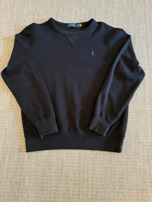 Svart stickad tröja Polo Ralph Lauren - Svart stickad tröja från Polo Ralph Lauren med rund halsringning och diskret broderad logga på bröstet. Tröjan har långa ärmar och ribbade muddar vid ärmslut och nederkant. Perfekt för en stilren och klassisk look.