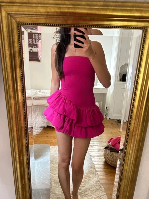 Axelbandslös rosa volangklänning - Säljer en superfin axelbandslös kort klänning i stark rosa färg. Klänningen har en tight överdel och två lager volanger i kjolen som ger en riktigt snygg och lekfull look. Perfekt för sommarens alla tillfällen!