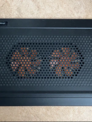 Svart laptopkylare med dubbla fläktar - Säljer en svart laptopkylare med två stora orange fläktar synliga genom ett perforerat metallgaller. Perfekt för att hålla datorn sval under gaming eller plugg. Stilren design och passar de flesta laptops.