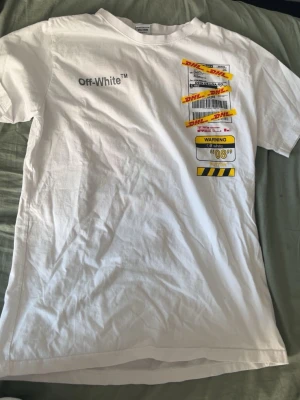 Vit Off-White t-shirt med DHL-tryck - Vit t-shirt från Off-White med DHL-inspirerat tryck och tecknad figur på ryggen. Framsidan har Off-White-logga och flera etikettmotiv. T-shirten är kortärmad och har en avslappnad passform, perfekt för en streetwear-look.