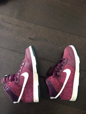 Vinröda Nike Dunk High sneakers limited edition - Säljer ett par vinröda Nike Dunk High sneakers limeted edition med vit sula och vit swoosh. Skorna har en mix av mocka och läderdetaljer med leopard aktigt möster samt klassisk hög siluett. Snygga detaljer och perforeringar på tån för extra stilpoäng. Köpta i new york för 9 år sedan men i gely nytt skick andvända 1 gång passade dock inte så ee har bara legat på en hyla och ska nu säljas.