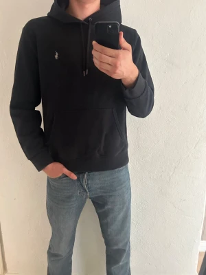 Svart hoodie från Polo Ralph Lauren - Säljer en svart hoodie från Polo Ralph Lauren med klassisk vit logga på bröstet. Hoodien har dragsko i huvan, stor magficka och ribbade muddar vid ärmar och nederkant. Perfekt för en chill och stilren look. Storlek L men funkar nog som M