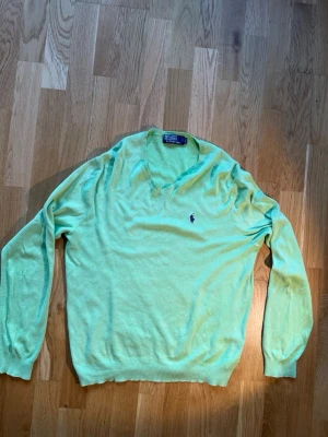 Grön stickad tröja från Polo Ralph Lauren - Snygg ljusgrön stickad tröja från Polo Ralph Lauren med V-ringning och klassisk broderad logga i mörkblått på bröstet. Tröjan har långa ärmar och ribbade muddar vid ärmslut och nederkant. Perfekt för lager på lager och stilren look.