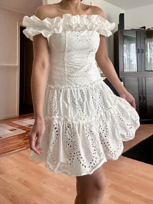 Vit offshoulder kort klänning med volang - Superfin vit kort klänning med offshoulder och volangkant upptill. Klänningen har broderade detaljer och är tillverkad i bomull. Perfekt för sommarens alla tillfällen och ger en romantisk vibe med sin luftiga kjol och feminina passform.