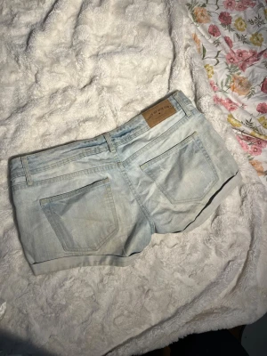 Lågmidjade shorts med slitningar - Säljer ett par ljusblå denimshorts med slitna detaljer och uppvikta benslut. Shortsen har klassiska fem fickor, bälteshällor och knappgylf. Perfekta för varma dagar och ger en avslappnad look.