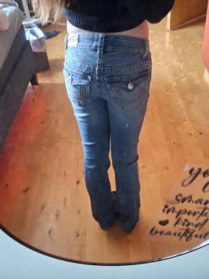 Blå bootcut jeans med fickdetaljer - Säljer ett par blå bootcut jeans med slitningar och dekorativa fickor bak med knappar. Jeansen har normalhög midja och klassisk femficksmodell. Perfekta för dig som gillar en avslappnad men ändå trendig stil.💕