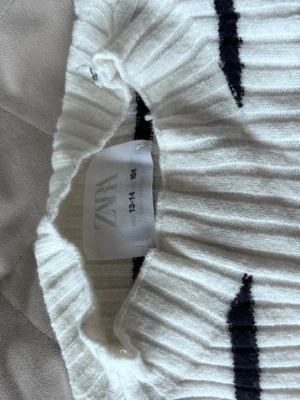 Vit ribbstickad tröja från Zara - Vit ribbstickad tröja från Zara med svarta horisontella ränder. Tröjan har långa ärmar och en högre krage. Materialet ser mjukt och stretchigt ut, perfekt för en avslappnad och trendig look.