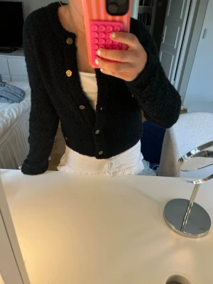 Svart fluffig kofta med guldknappar - Mysig svart kofta med fluffig textur och långa ärmar. Koftan har flera dekorativa guldknappar framtill och en något croppad passform. Perfekt att slänga över ett linne eller t-shirt för en chill look.