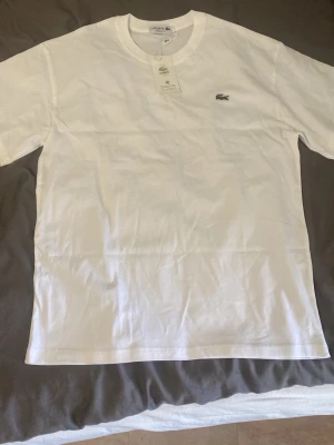 Vit t-shirt från Lacoste - Klassisk vit t-shirt från Lacoste med den ikoniska gröna krokodilloggan broderad på bröstet. T-shirten har rund halsringning och korta ärmar. Tillverkad i mjuk bomull för en skön känsla och enkel stil.