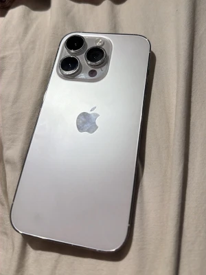 Vit iphone 14pro - Snygg vit iPhone med stilren design och tre stora kameralinser på baksidan. Telefonen har en blank yta i glas och den klassiska Apple-loggan i mitten. Perfekt för dig som gillar modern teknik och vill ha en mobil med grym kamera. Några repor och någon spricka men annars bra