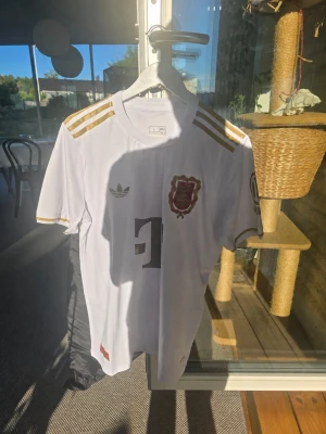 Vit FC Bayern München jubileumströja - Snygg vit matchtröja från Adidas för FC Bayern München, jubileumsmodell 125 års jubileum. Med guldfärgade detaljer och klubbmärke med '125' broderat på bröstet. Tröjan har korta ärmar och tre guldiga ränder på axlarna. Är i Extremt bra kvalité och alla emblem o sånt är påsydd. Perfekt för dig som älskar fotboll och samlar på unika matchtröjor.