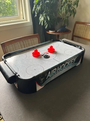 Bordmodell airhockeyspel - Säljer ett kompakt airhockeyspel i bordmodell med vit spelplan, svarta kanter och tydliga markeringar. Två röda handtag och en svart puck ingår. Perfekt för att köra snabba matcher hemma med vänner. Spelet är gjort i plast och har poängräknare på båda sidor.