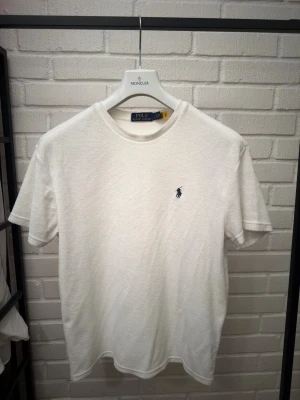 Vit t-shirt från Polo Ralph Lauren - Klassisk vit t-shirt från Polo Ralph Lauren med den ikoniska broderade loggan i marinblått på bröstet. T-shirten har rund halsringning och korta ärmar. Tillverkad i mjuk bomull för en skön känsla och enkel stil.