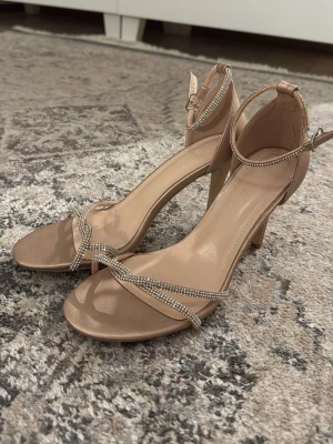 Beige pumps med smala glittriga remmar - Säljer ett par snygga beige pumps med smala, glittriga remmar och öppen tå. Skorna har en elegant ankelrem med spänne och en smal klack som ger en stilren look. Perfekta för att lyfta vilken outfit som helst med lite extra bling.