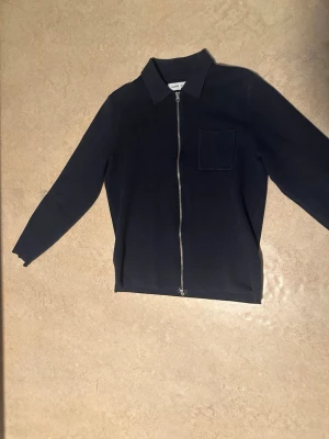 Svart overshirt med dragkedja Samsøe - Stilren svart overshirt från Samsøe Samsøe med rak passform, klassisk krage och silverfärgad dragkedja framtill. En bröstficka på vänster sida och mjukt tyg som känns skönt mot huden. Perfekt lager på lager-plagg för en clean look.