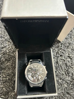 Emporio Armani klocka - Säljer en stilren klocka från Emporio Armani. Asken är vit på utsidan med logga och har en elegant silverfärgad innerbox med präglad logotyp. Mycket bra skick men batteriet behöver bytas 