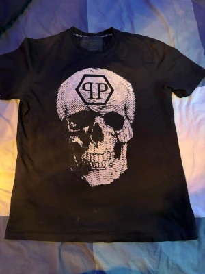 Svart t-shirt med dödskalle - Snygg svart t-shirt med ett stort dödskalletryck i vitt och PP-logga framtill. Klassisk rund hals och korta ärmar. Perfekt för dig som gillar edgy streetstyle och vill sticka ut. T- shirten är perfekt för att göra dig att sticka ut i allmänheten.