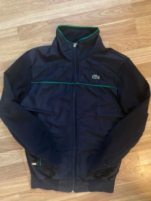 Lacoste hoodie  - Snygg marinblå vindjacka från Lacoste med dragkedja framtill, hög krage och gröna detaljer längs bröstet och insidan av kragen. Klassisk krokodil-logga på vänster bröst. Perfekt för dig som gillar sportig och stilren look.
