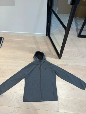  Under Armour  - Snygg grå vindjacka från Under Armour med minimalistisk design och huva. Jackan har dragkedja framtill, två sidofickor och diskret logga på bröstet. Perfekt för blåsiga dagar och enkel att matcha med olika outfits.