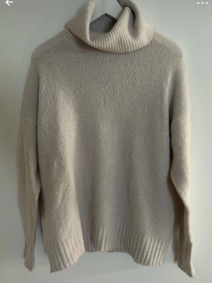 Beige polotröja i stickat material - Mysig beige stickad polotröja med bred krage och avslappnad passform. Tröjan har långa ärmar och ribbade muddar vid ärmslut och nederkant. Perfekt för kyliga dagar och enkel att matcha med olika outfits.