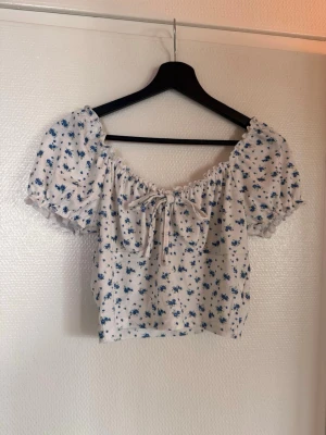 Vit blommig croppad blus med puffärm - Supersöt vit croppad blus med blått blommönster och puffiga korta ärmar. Toppen har en söt knytning framtill och elastisk kant vid axlarna som ger en romantisk vibe. Perfekt för varma dagar och somriga outfits.