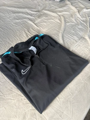 Svart Nike träningströja med dragkedja - Snygg svart träningströja från Nike med halv dragkedja framtill och turkosa detaljer på axlarna. Tillverkad i ett lätt och funktionellt material som passar perfekt för träning eller uppvärmning. Nike-logga broderad på bröstet.