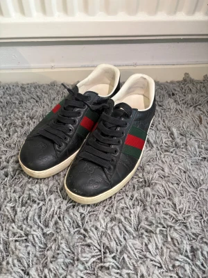 Svarta sneakers med röd/grön rand - Snygga svarta sneakers i läder med klassiska gröna och röda ränder på sidan. Skorna har svarta skosnören, vit sula och diskret mönster i lädret. Perfekta för dig som vill ha en stilren och trendig look. Priset är inte hugget i sten vid snabb affär, skriv gärna vid minsta lilla fundering!
