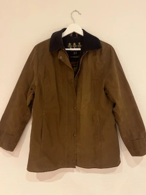 Brun vaxad rock från Barbour - Klassisk brun vaxad rock från Barbour med mörkbrun manchesterkrage och rutigt innerfoder. Rocken har tryckknappar, dragkedja och raka ärmar. Perfekt för dig som gillar tidlös brittisk stil och funktionella material.