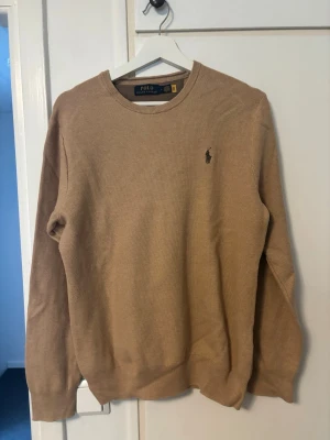 Beige tröja från Polo Ralph Lauren - Snygg beige tröja från Polo Ralph Lauren med rund halsringning och diskret broderad logga på bröstet. Tröjan har lång ärm och ribbade muddar vid ärmslut och nederkant. Perfekt för en clean och klassisk stil.