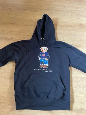Svart hoodie Polo Ralph Lauren M - Svart hoodie från Polo Ralph Lauren med den ikoniska Polo Bear-figuren på bröstet. Hoodien har dragsko i huvan, stor magficka och ribbade muddar. Materialet är mjukt och bekvämt, perfekt för en avslappnad stil.