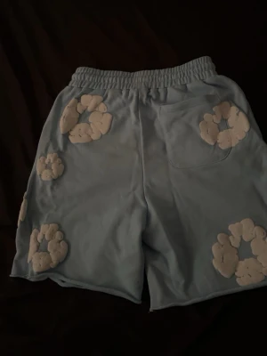 Ljusblå shorts med vita blommor - Unika ljusblå shorts med elastisk midja och snörning. Dekorerade med stora vita blommor i ett mjukt material över hela plagget. Perfekta för dig som vill sticka ut med en lekfull och trendig stil.