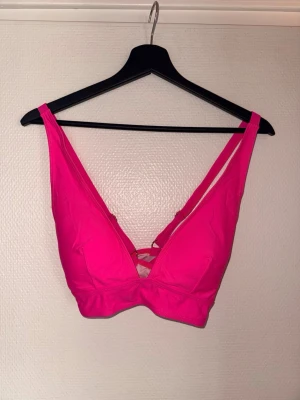 Rosa bikiniöverdel med djup urringning - Säljer en snygg bikiniöverdel i starkt rosa färg. Modellen har djup urringning och breda axelband för extra stöd. Materialet känns mjukt och stretchigt, perfekt för strandhäng eller poolen. Detalj med korsade band framtill ger en trendig touch.