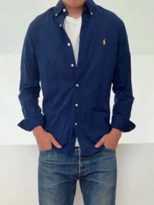 Mörkblå skjorta från Ralph Lauren - Klassisk mörkblå skjorta från Ralph Lauren med button-down krage och vita knappar. Skjortan har lång ärm och den ikoniska gula loggan broderad på bröstet. Tillverkad i bomull för en skön och stilren look.