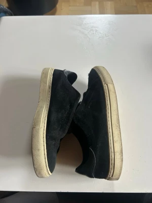 Axel Arigato sneakers - Säljer ett par svarta sneakers i mocka med vit sula och dragkedja på ovansidan. Skorna har en låg profil och klassisk design som passar till det mesta. Insidan är fodrad med svart material och sulan har en diskret struktur. Det finns en rispa på  ena skon och sedan så har lite av sömmen släppt också, men det går lätt att åtgärda