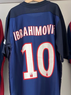 PSG Ibrahimović matchtröja blå - Snygg blå Paris Saint-Germain matchtröja med Ibrahimović och nummer 10 på ryggen. Tröjan har röda och vita detaljer, PSG-logga på bröstet och Fly Emirates som sponsor. Tillverkad i lätt och ventilerande material, perfekt för fotboll eller samling.
