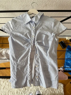 Blårandig kortärmad skjorta från Gina Tricot - Snygg kortärmad skjorta med blå och vita ränder från Gina Tricot i storlek XS. Skjortan har klassisk krage, knappar framtill och ett knytband i ryggen för extra passform. Är knappt använd, den andra knappen är lite lös då den va så när jag köpte den men det är inget som syns och går att knäppa ändå😊