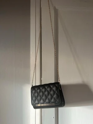 Svart quiltad kuvertväska med kedja - Snygg svart kuvertväska med quiltat mönster och guldfärgad kedja som axelrem. Väskan har en elegant gulddetalj på framsidan och är tillverkad i konstläder. Perfekt för att lyfta din outfit med en stilren touch.