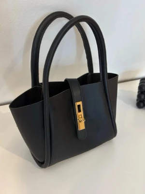 Svart handväska med gulddetalj - Stilren svart handväska i slät konstläder med två rundade handtag och en snygg guldfärgad låsdetalj framtill. Väskan har en minimalistisk design och rymlig insida, perfekt för att bära det viktigaste. Passar till många olika outfits.