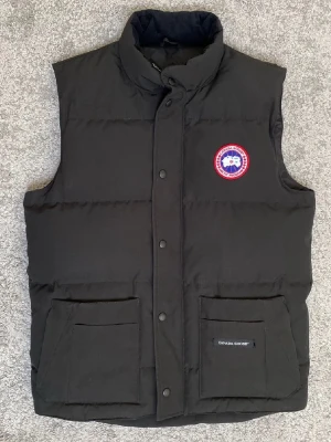 Svart dunväst från Canada Goose - Snygg svart dunväst från Canada Goose med klassisk logga på bröstet. Västen har hög krage, två stora fickor framtill och stängs med tryckknappar. Perfekt för lager-på-lager och håller dig varm under kyliga dagar.