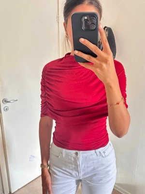 Röd draperad topp med rynkade ärmar - Snygg röd topp med draperad effekt och rynkade detaljer längs ärmarna. Modellen är tight och har korta ärmar. Perfekt för dig som vill ha en trendig och färgstark look. Tillverkad i mjukt och stretchigt material.