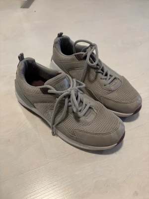 Grå mesh sneakers med vita sulor - Snygga grå sneakers med ovandel i mesh och syntetmaterial. Skorna har vita sulor med vinröda detaljer undertill och på plösen. Klassisk snörning och låg profil gör dem perfekta för en avslappnad stil.