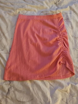 Rosa kortkjol med rynkdetalj - Säljer en rosa kortkjol i ett ribbat material med snygg rynkdetalj på sidan. Kjolen har en lätt asymmetrisk nederkant och är stretchig för en bekväm passform. Perfekt för dig som vill ha en trendig och färgglad look.