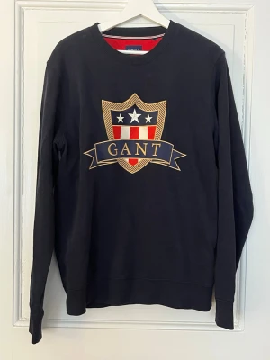 Blå sweatshirt med GANT-logga - Stilren mörkblå sweatshirt från GANT med stort broderat sköldmärke och text på bröstet. Tröjan har rund halsringning, långa ärmar och ribbade muddar. Perfekt för en avslappnad och trendig look. Nypris 1000kr