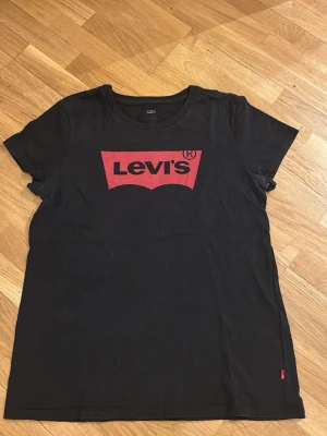 Svart Levi's t-shirt med röd logga - Säljer en svart t-shirt från Levi's med den klassiska röda loggan tryckt på bröstet. T-shirten har rund halsringning och korta ärmar. Tillverkad i mjuk bomull som är skön att bära. Perfekt till jeans eller shorts för en avslappnad look.