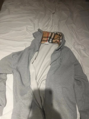 Grå hoodie  Burberry med mönstrad huva - Snygg grå zip-hoodie från Burberry med den klassiska rutiga Burberry-mönstrade huvan. Hoodien har dragkedja, fickor på sidorna och en diskret Burberry-logga på bröstet. Perfekt för dig som vill ha en stilren men ändå lyxig look.