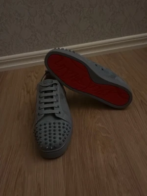Grå sneakers med nitar och röd sula - Säljer ett par grå sneakers med coola nitar över tån och klassisk röd sula. Skorna har snörning och en stilren design som sticker ut. Perfekta för dig som vill ha något unikt och edgy i din skogarderob.
